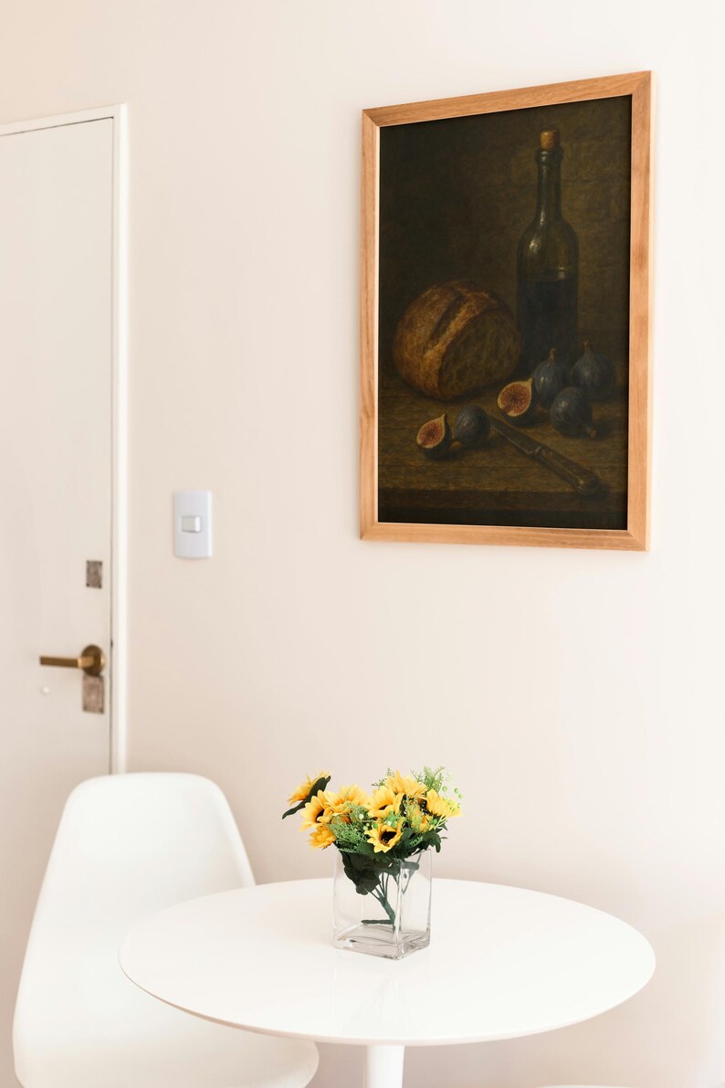 Pode incluir: Uma pintura emoldurada de natureza morta retrata um p&atilde;o, figos, uma faca e uma garrafa de vinho. Um pequeno vaso de girass&oacute;is est&aacute; em uma mesa branca em frente a uma cadeira branca.
