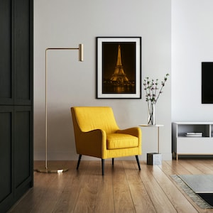 May include: A modern living room featuring a yellow armchair, a gold floor lamp, and a framed photograph of the Eiffel Tower at night. The room has hardwood floors and a minimalist design, with a white side table and a black cabinet.