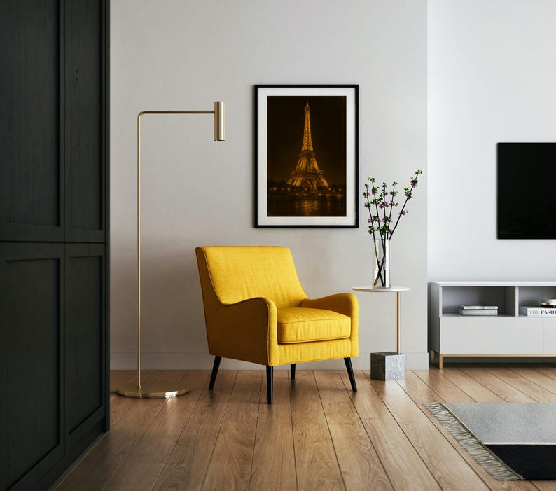 May include: A modern living room featuring a yellow armchair, a gold floor lamp, and a framed photograph of the Eiffel Tower at night. The room has hardwood floors and a minimalist design, with a white side table and a black cabinet.
