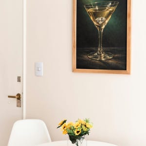 Puede incluir: Un cuadro enmarcado de una copa de martini con un diamante en la bebida cuelga de una pared. Una mesa peque&ntilde;a sostiene un jarr&oacute;n con girasoles. Una silla blanca est&aacute; cerca de la mesa. La habitaci&oacute;n tiene una paleta de colores neutros.