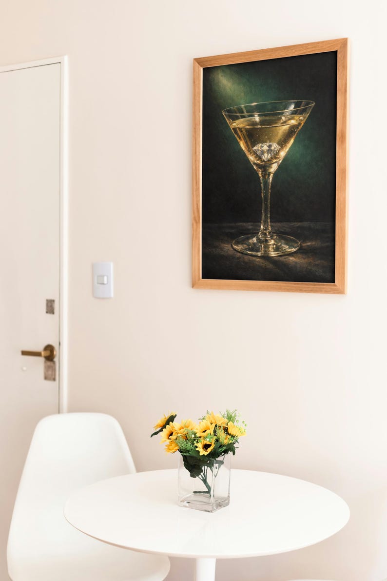 Puede incluir: Un cuadro enmarcado de una copa de martini con un diamante en la bebida cuelga de una pared. Una mesa peque&ntilde;a sostiene un jarr&oacute;n con girasoles. Una silla blanca est&aacute; cerca de la mesa. La habitaci&oacute;n tiene una paleta de colores neutros.