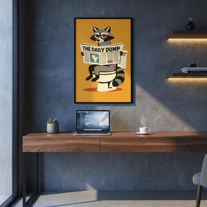 Op de afbeelding: Een ingelijste kunstprint met een wasbeer met een bril die de krant "THE DAILY DUMP" leest op een toilet. Het kunstwerk heeft een gele achtergrond en staat boven een houten bureau met een laptop en een kleine plant.