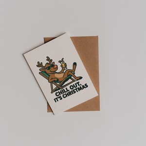 Pu&ograve; includere: Un biglietto di Natale con una renna dei cartoni animati con occhiali da sole, che si rilassa su una sedia a sdraio e tiene in mano un cocktail. Il biglietto riporta la scritta "CHILL OUT, IT'S CHRISTMAS" ed &egrave; posto su una busta marrone.