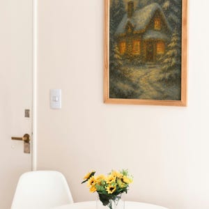 K&ouml;nnte beinhalten: Ein gerahmtes Gem&auml;lde eines schneebedeckten Hauses mit beleuchteten Fenstern h&auml;ngt an einer Wand. Ein kleiner wei&szlig;er Tisch tr&auml;gt eine klare Glasvase mit gelben Sonnenblumen. Ein wei&szlig;er Stuhl steht neben dem Tisch. Der Raum hat eine neutrale Farbpalette.