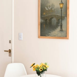 May include: A framed painting depicts a snowy bridge scene with a lamppost. A vase of sunflowers sits on a white table. A white chair is next to the table. The painting has a wooden frame. The room has a white door and walls.