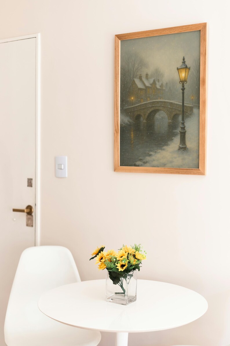 May include: A framed painting depicts a snowy bridge scene with a lamppost. A vase of sunflowers sits on a white table. A white chair is next to the table. The painting has a wooden frame. The room has a white door and walls.