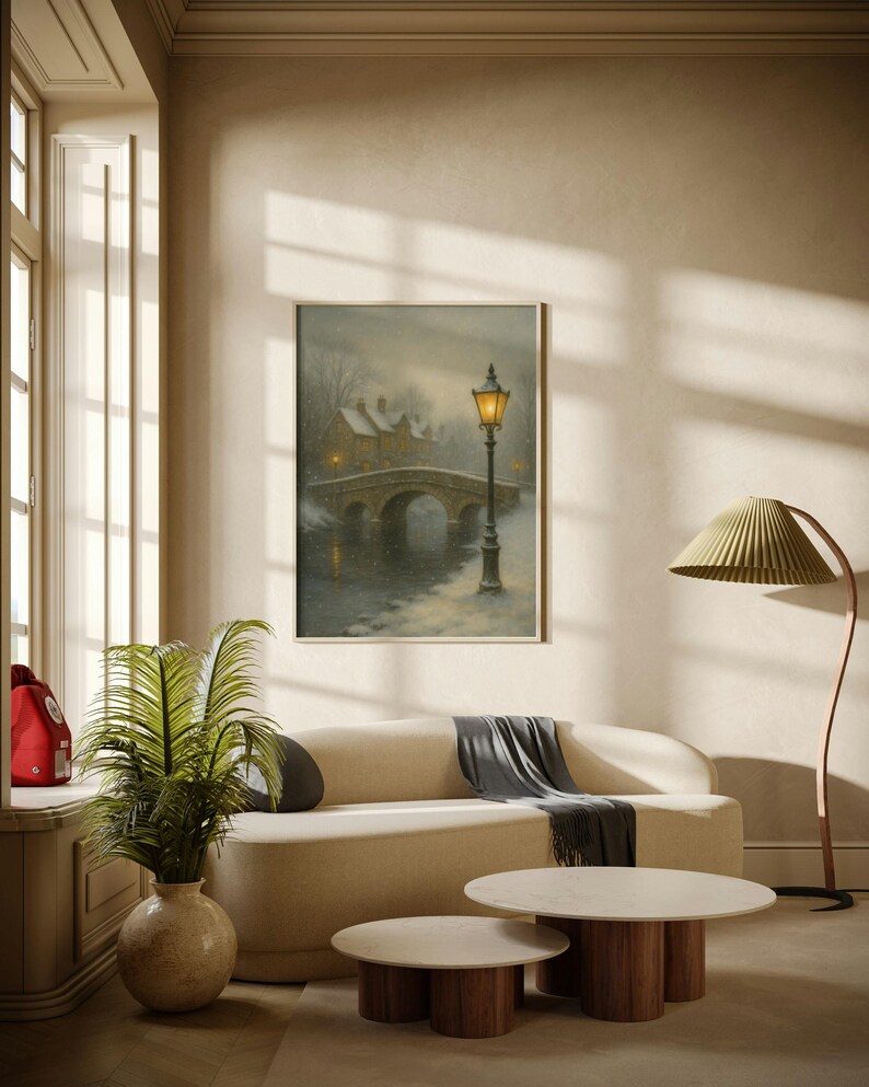 May include: A framed painting depicts a winter scene with a stone bridge, a lamppost, and snow-covered buildings. The artwork hangs above a cream-colored sofa with a dark gray throw. Two round coffee tables with wooden bases and marble tops sit in front of the sofa.