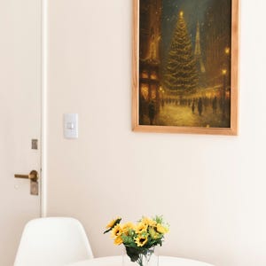 May include: A framed print of a Christmas scene in a city, with a large decorated tree and the Eiffel Tower in the background. A small bouquet of sunflowers sits on a white table in front of a white chair.