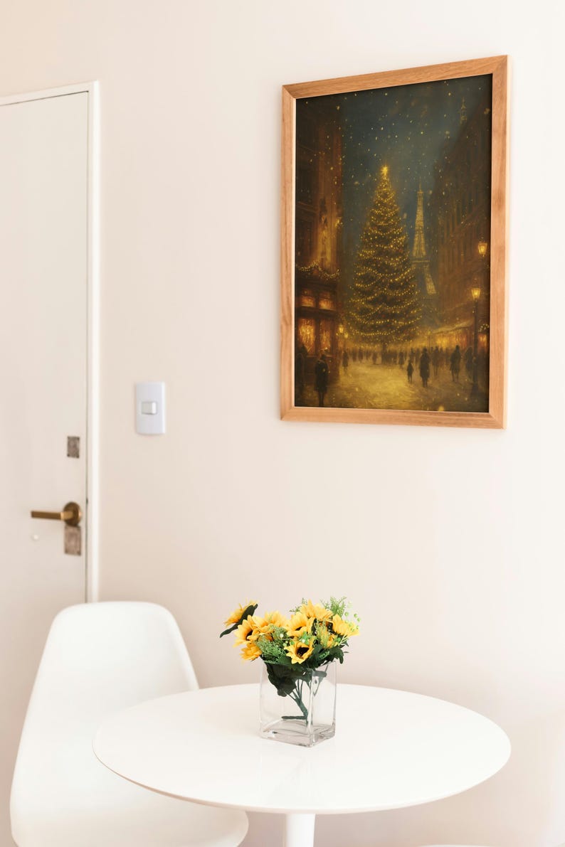 May include: A framed print of a Christmas scene in a city, with a large decorated tree and the Eiffel Tower in the background. A small bouquet of sunflowers sits on a white table in front of a white chair.