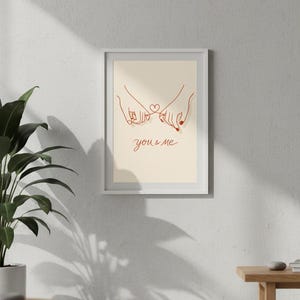 Puede incluir: Impresi&oacute;n art&iacute;stica enmarcada con un dibujo lineal minimalista de dos manos entrelazando los me&ntilde;iques, con un coraz&oacute;n y las palabras "you & me" en rojo. La obra de arte se muestra en un marco blanco.