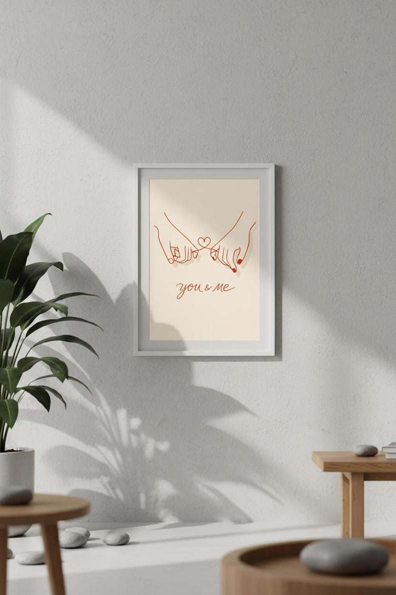Puede incluir: Impresi&oacute;n art&iacute;stica enmarcada con un dibujo lineal minimalista de dos manos entrelazando los me&ntilde;iques, con un coraz&oacute;n y las palabras "you & me" en rojo. La obra de arte se muestra en un marco blanco.