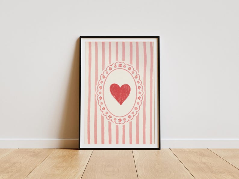 Op de afbeelding: Ingelijste kunstdruk met een rood hart in een decoratief ovaal op een gestreepte achtergrond. Het kunstwerk staat in een zwarte lijst en leunt tegen een witte muur. De achtergrond heeft verticale roze strepen.