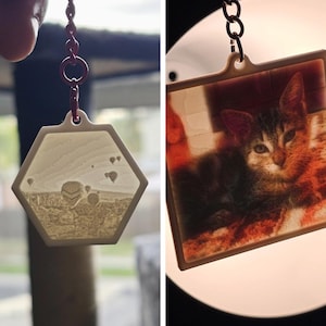 May include: Two keychains are shown. One is a cream-colored, hexagonal keychain with a landscape of hot air balloons. The other is a rectangular keychain with a photo of a kitten. Both keychains have a red chain.