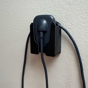 Op de afbeelding: Een donkerblauwe oplaadadapter en kabel worden in een zwarte wandhouder gehouden. De adapter is vierkant met een afgeronde bovenkant en één poort. De kabel loopt naar beneden vanaf de adapter.