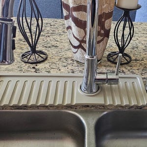 May include: A stainless steel kitchen faucet with a pull-down sprayer is installed over a double sink. A light gray, ribbed sink mat sits beneath the faucet. Decorative items include a vase and candle holders.