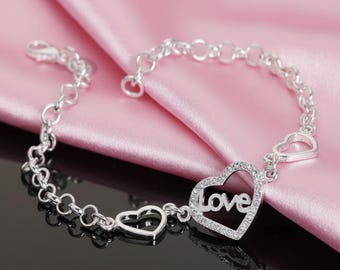 Pode incluir: Pulseira prateada com um pendente em forma de coração com a palavra "Love" no centro. A pulseira tem um design de corrente delicada com detalhes adicionais em forma de coração. Apresentada em tecido de cetim rosa.