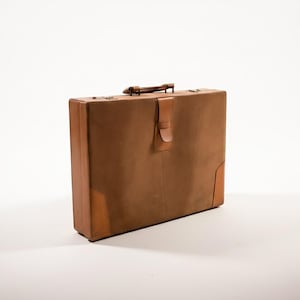 May include: A vintage brown leather briefcase with a handle and a clasp closure. The briefcase has reinforced corners and a flap closure. The handle is a darker brown color. The briefcase is set against a white background.