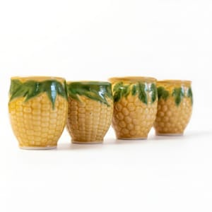 Op de afbeelding: Vier keramische shotglaasjes in de vorm van maïskolven. De glazen zijn geel met een textuur en groene, bladerrijke toppen. De shotglaasjes zijn in een rij op een witte achtergrond geplaatst.