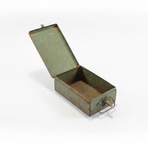 Puede incluir: Una caja metálica rectangular verde oliva abierta, con tapa abisagrada. La caja tiene un asa en la parte delantera y muestra signos de desgaste y óxido. La tapa está ligeramente entreabierta, revelando el interior. La caja probablemente se utiliza para almacenamiento.
