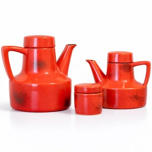 Peut inclure: Ensemble de trois articles de cuisine en céramique rouge : deux théières et un petit pot. Les théières ont des couvercles et des poignées. Le pot a un couvercle. Les articles ont une finition brillante avec des accents brun foncé. Fond blanc.