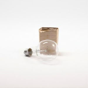 Puede incluir: Una bombilla de vidrio transparente con base plateada, junto a una pequeña caja de cartón abierta. El filamento es visible dentro de la bombilla. El fondo es blanco.