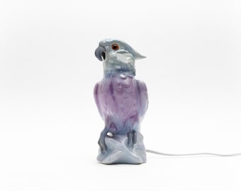 May include: A ceramic parrot figurine in shades of purple, blue, and gray. The bird is perched on a rock-like base, with a detailed beak and eye. A white electrical cord extends from the base. The figurine is a decorative item.