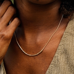 May include: A delicate gold necklace with small, evenly spaced beads. The necklace is worn on a person's neck, with the clasp hidden from view. The person is wearing a textured, olive-green top.