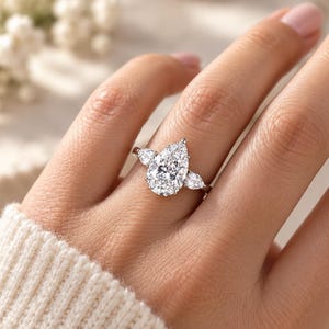 May include: A silver-toned ring featuring a large pear-cut center stone flanked by two smaller pear-cut stones on a thin band. The setting is metallic and polished, showcasing clear, faceted stones that reflect light across the surface.