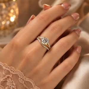 May include: A gold ring featuring a large, round, brilliant-cut center stone held by four prongs, with a split shank design encrusted with small pavé diamonds. The band curves slightly to accommodate the center setting, creating a layered look.