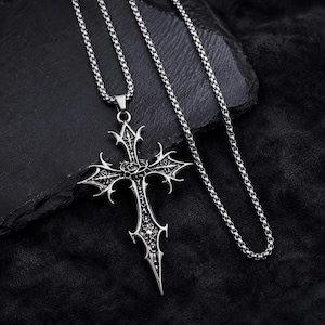 May include: A silver-toned metal cross pendant featuring intricate filigree detailing and a central rose motif, suspended from a matching box chain necklace. The cross has sharp, pointed edges and a polished finish with darkened recessed areas.