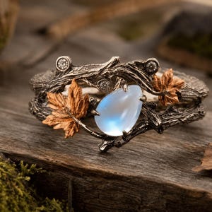 May include: A ring featuring a dark, textured band designed to resemble intertwined tree branches. The ring is adorned with a central pear-shaped, iridescent moonstone, two small copper-colored maple leaves, and three small, clear round stones.