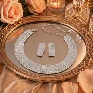 May include: A silver-toned jewelry set consisting of a wide, flexible choker necklace and a pair of matching rectangular drop earrings. Both pieces are densely encrusted with small, clear, reflective rhinestones that create a sparkling effect.
