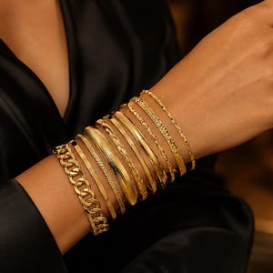 May include: A stack of ten gold-toned bracelets featuring various textures and designs, including a thick curb chain, smooth polished bands, and intricate twisted patterns. The collection is worn together on the wrist, showcasing a layered aesthetic.