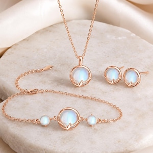 May include: A rose gold jewelry set consisting of a necklace, a pair of stud earrings, and a bracelet. Each piece features a round, iridescent moonstone cabochon set within a delicate metal frame that resembles a small, stylized leaf or branch.