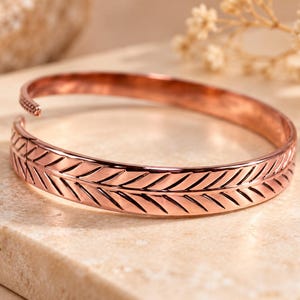May include: A copper-toned open cuff bracelet featuring a continuous engraved feather or leaf pattern along the outer surface. The metal band has a polished finish with dark recessed lines highlighting the detailed chevron-like texture.