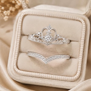 May include: A silver-toned ring set consisting of two pieces. The top ring features a crown-shaped design with a central round stone and smaller accent stones. The bottom ring is a V-shaped band encrusted with numerous tiny clear stones.