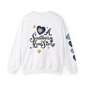 May include: White sweatshirt with the text "A Southern Love Story" in a script font, with heart graphics in blue and gold. The sleeves have additional heart graphics with text.