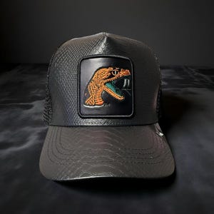 May include: A black baseball cap with a textured snakeskin pattern on the front panels and brim. The cap features a central rectangular patch displaying an embroidered orange and teal snake head with visible fangs against a black background.
