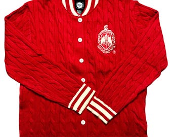 Puede incluir: Chaqueta universitaria roja de punto trenzado con rayas blancas en el cuello, los puños y el dobladillo. La chaqueta tiene botones blancos y un emblema bordado en el pecho. El emblema incluye texto y un diseño de escudo.