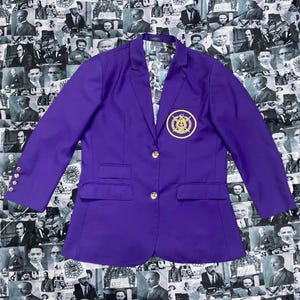 Peut inclure: Un blazer violet vif avec des boutons dorés et un écusson assorti sur la poitrine. Le blazer a un design classique avec deux poches avant et une doublure à motifs. Le fond présente un collage de portraits en noir et blanc.