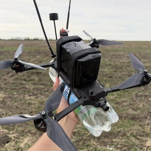 Puede incluir: Un dron negro y gris con cuatro hélices, una funda de almacenamiento negra y múltiples antenas. El dron se sostiene sobre un campo con una botella de plástico transparente adjunta.