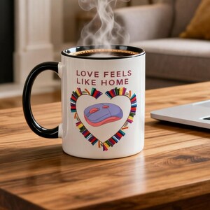 May include: A white coffee mug with a black handle and rim, featuring the text "LOVE FEELS LIKE HOME." A heart-shaped design includes a computer mouse and books. Steam rises from the coffee, suggesting it is hot.