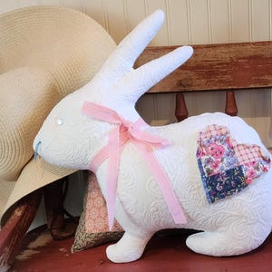 May include: A white quilted bunny-shaped pillow with a pink ribbon bow and a patchwork heart detail. A large straw hat rests behind the bunny. The bunny is sitting on a red bench.