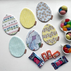 May include: A set of six colorful fabric coasters with various patterns, including floral, geometric, and abstract designs. The coasters are arranged on a white surface with small bowls of candy and chocolate eggs.