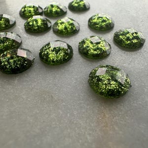 May include: Close-up of several green glittery cabochons with faceted edges. The cabochons are oval-shaped and have a sparkling, textured appearance. They are scattered on a light gray surface, creating a shimmering effect. These could be used for jewelry making or craft projects.