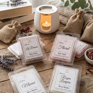 Puede incluir: Fundidos de cera de aromaterapia en envases transparentes, con etiquetas "La Dama", "Floral", "Monoi" y "Ropa Limpia". Un quemador de aceite de cerámica blanca con una vela encendida sobre una superficie de madera.