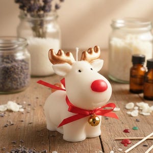Puede incluir: Una vela blanca con forma de reno con cuernos dorados y una nariz roja, adornada con una cinta roja y una pequeña campana dorada. La vela está sobre una superficie de madera, con frascos de lavanda y otros ingredientes al fondo.