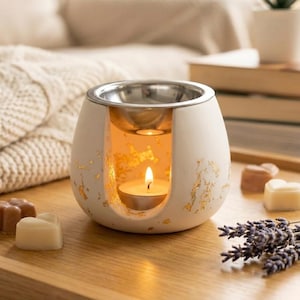 Könnte beinhalten: Cremefarbener Diffusor für ätherische Öle mit silberfarbener Oberseite und Goldblattakzenten. Eine brennende Teelichtkerze leuchtet im Inneren des Diffusors. Der Diffusor steht auf einem Holztablett mit herzförmigen Wachsschmelzen und Lavendelzweigen.