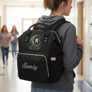 Peut inclure: Sac à dos noir orné d'un croissant de lune et d'une étoile, avec des accents de fougères et le nom "Everly" en écriture turquoise. Le sac possède plusieurs compartiments, une poignée supérieure et des bretelles réglables. Le sac est porté dans un couloir d'école.