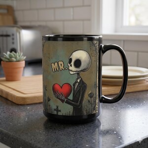 May include: Black ceramic mug featuring a skeleton holding a red heart, with the word "MR." above. The design has a vintage, distressed look with a graveyard scene. The mug has a black handle and interior.